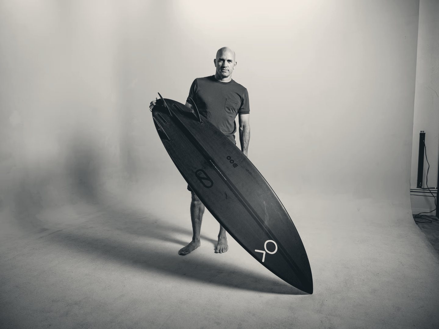 Kelly Slater standing in a studio holding a blacked-out surfboard